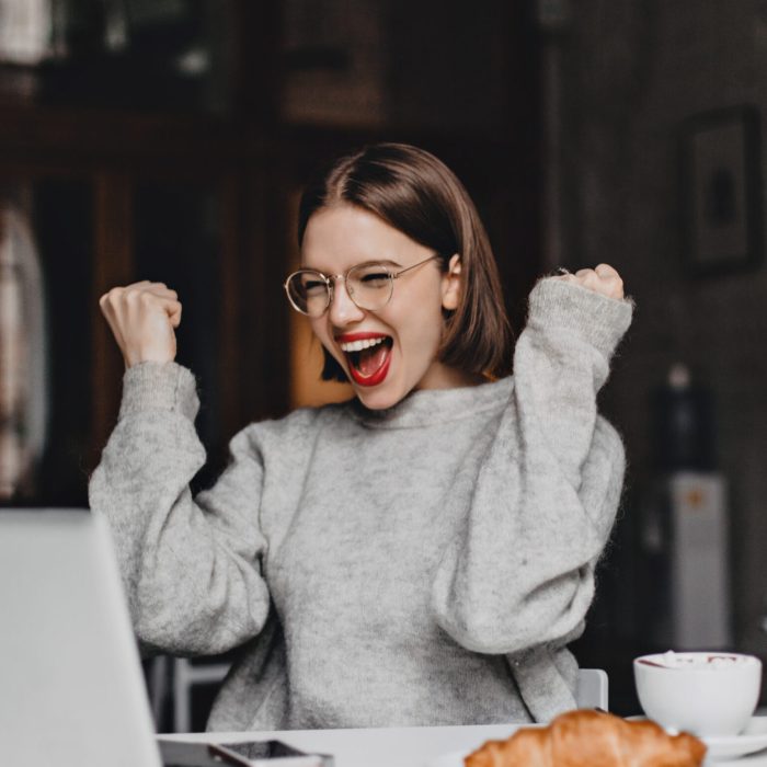 Happy woman in glasses makes winning gesture and sincerely rejoices. Lady with red lipstick dressed in gray sweater looking at laptop. Happy woman in glasses makes winning gesture and sincerely rejoices. Lady with red lipstick dressed in gray sweater looking at laptop
